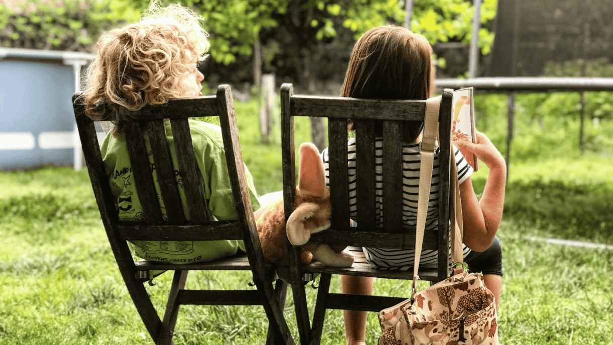 Zwei Kinder sitzen im Garten auf Stühlen und lesen ein Buch.