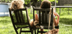 Zwei Kinder sitzen im Garten auf Stühlen und lesen ein Buch.